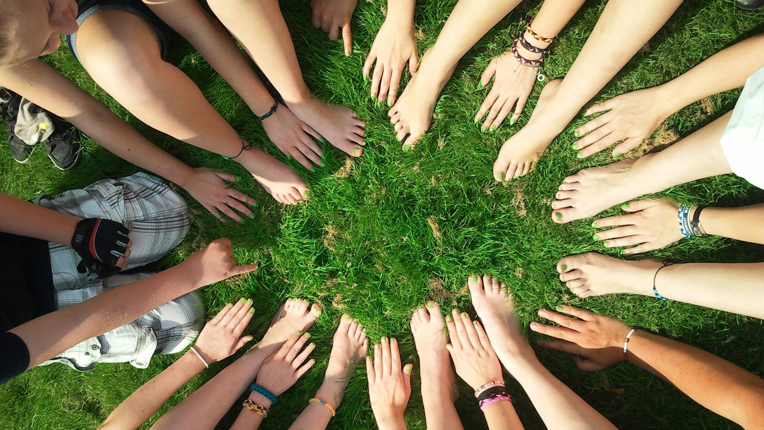 Group of friends forming a circle with their hands and feet on grass, symbolizing unity and friendship.